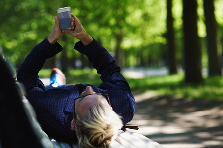 Blond male in glasses and checkered jacket lying on back on white park bench and looking at mobile phone in stretched out arms on sunny summer day on blurred backgroundの写真素材