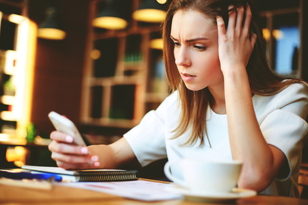 Puzzled young woman surfing smartphone and facing problems while working with papers and having coffee in cafeの写真素材