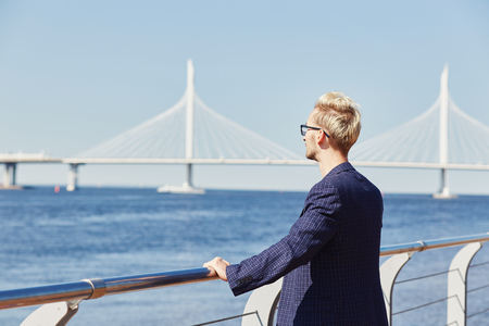 Back view of blond man in glasses and dark jacket enjoying view of water surface at summer leaning on metal handrail and looking away with suspension bridge on backgroundの写真素材