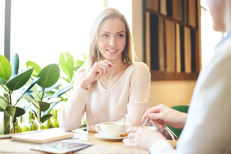 Young woman sitting at table with friend or business partner while having coffee meeting in daylightの写真素材