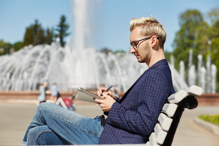 Side view of handsome blond male in glasses and dark checkered jacket sitting with crossed legs on park bench and concentrating on writing in notebook with sunlit fountain on blurred background at summerの写真素材