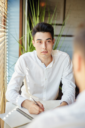 Young man in white shirt sitting with anonymous colleague having meeting and taking notes in notepadの写真素材