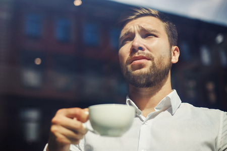 From below shot of elegant businessman behind window glass holding cup of coffee in sunlight looking awayの写真素材