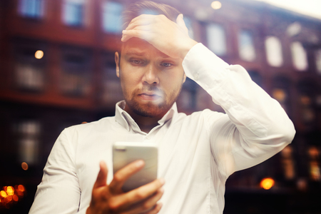 Shot through glass of stunned young businessman holding hand on forehead and watching phone in puzzlementの写真素材