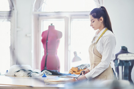 Side view of beautiful fashion designer standing near table and cutting cloth during workdayの写真素材