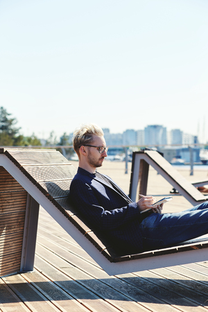 Side view of blond male in glasses and dark elegant jacket concentrating on writing in notebook sitting relaxed on lounge chair on embankment with wooden flooring at summerの写真素材