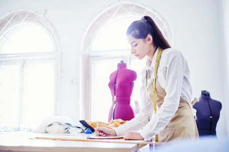 Side view of beautiful young woman in apron standing near table and drawing in notebook while working in light tailor workshopの写真素材