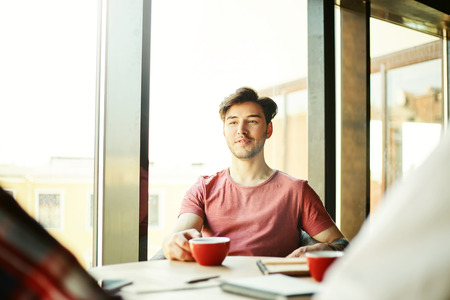 Young man in casual clothing sitting at cafe table and having coffee during meetingの写真素材