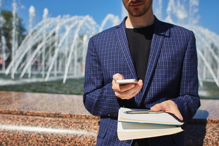 Crop view of male in dark checkered jacket holding notepad with pen and looking at mobile phone in hands with sunlit fountain on backgroundの写真素材