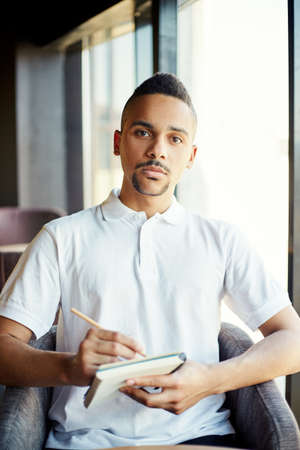 Portrait of young mixed race businessman in casual clothing looking at camera while making notes in his notebookの写真素材
