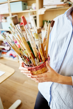 Unrecognizable young teacher holding basket with various brushes while standing in classroom in art schoolの写真素材