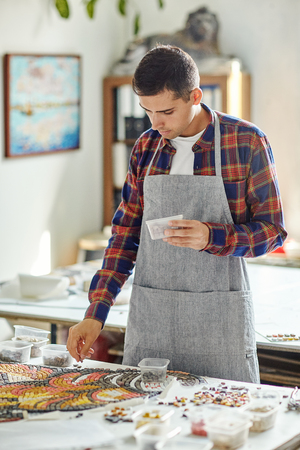 Casual man in gray apron laying out colorful mosaic on table creating beautiful piece of artの写真素材