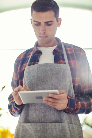 From below shot of young man in apron using tablet while standing in sunny workshop aloneの写真素材