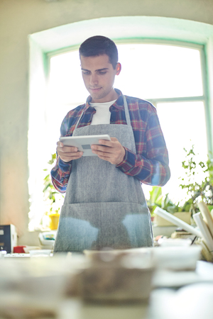 Relaxed young artisan in apron using tablet standing in workshop with mosaic art on tableの写真素材