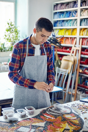 Young casual man composing multicolored mosaic details on table working in studioの写真素材