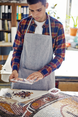Casual man in apron working with mosaic layout and using tablet to check designの写真素材