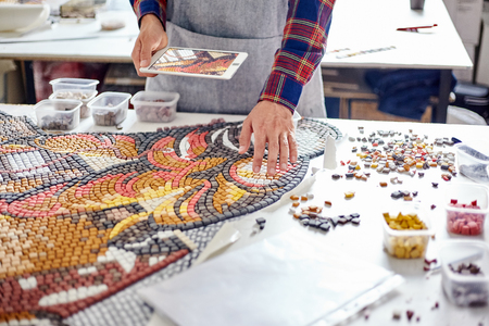 Faceless shot of man in apron standing at table in workshop and composing beautiful mosaic in daylightの写真素材