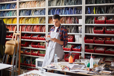 Modern man in apron standing in workshop with colorful ceramic pieces on shelves and using tabletの写真素材