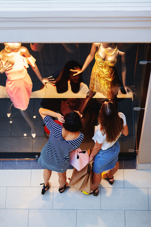 From above shot of girlfriends shopping in mall and looking at mannequins in stylish clothes behind window glassの写真素材
