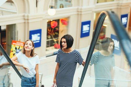 Young stylish women standing on moving stairs while spending time together in shopping centerの写真素材