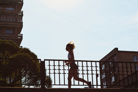 Side view of young sportswoman jogging near metal fence on city street during outdoor training on sunny dayの写真素材