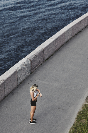 From above shot of young lady in sportswear looking on fitness tracker while standing on asphalt path near river during trainingの写真素材
