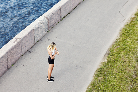 Side view of anonymous woman in sportswear standing on asphalt path and looking at fitness tracker during outdoor training on embankmentの写真素材