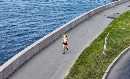 From above shot of young lady in sportswear running on asphalt path between green lawn and river during outdoor training on embankmentの写真素材