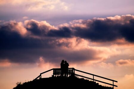 Silhouette of a couple standing on a hill near a bench close to a fence and admiring a dramatic...
