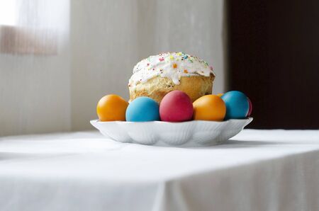 White plate with a glazed easter cake and painted eggs standing on a table covered with a white...