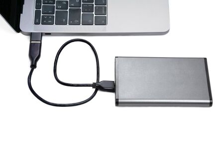 Dark grey portable hard disk drive connected to a laptop computer via adaptor isolated on white.