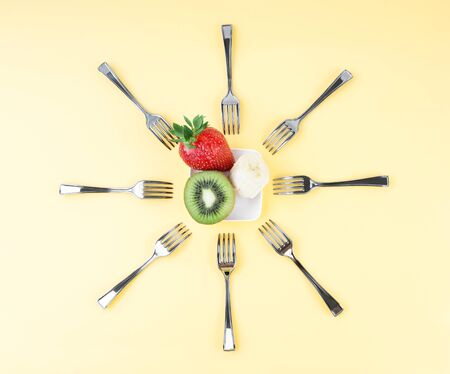 Creative composition in the form of the sun made of forks arranged around a tiny plate with a piece of banana, half of a kiwi and a strawberry. Top refreshing and energy boost fruits snack concept.の写真素材