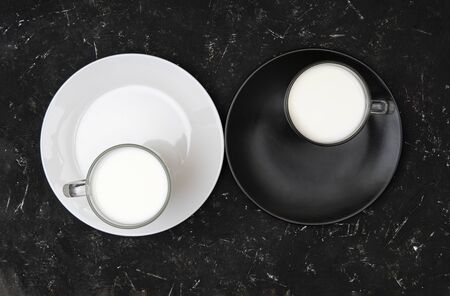 Minimalist flat lay of two mugs of milk standing on black and white plates, resembling the yin yang concept on blackの写真素材