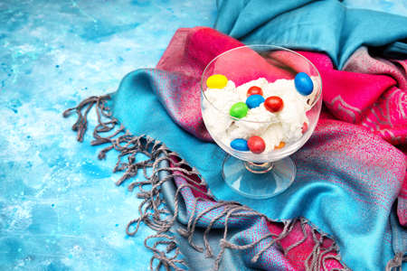 Ice cream with multicolor candies in a glass bowl standing on a colorful cloth and light blue...