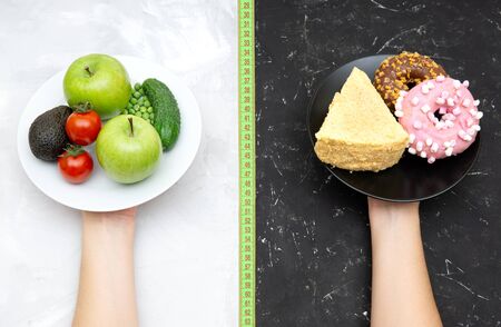 Female hands holding white plate with vegetables and fruits and black plate with donuts and a cake on contrast background divided with a measuring tape. Choosing between healthy and junk food concept.の写真素材