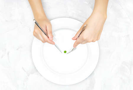 Female hands trying to catch a small green pea on a large white plate with knife and fork. Eating disorder concept.の写真素材