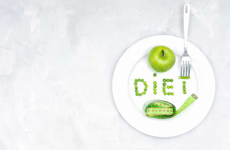 Word DIET made of garden peas on a large white plate with a fresh green apple, a soft tape measure and a fork on a concrete table. Healthy lifestyle, weight loss and dieting concept.の写真素材