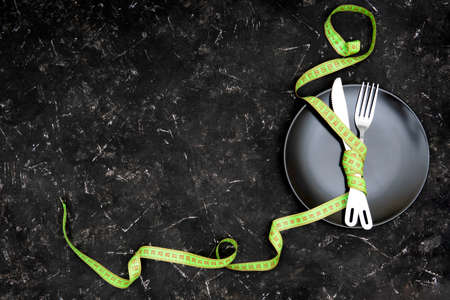 A fork and a knife tied up with a green measuring tape on a black plate on a textured black table....
