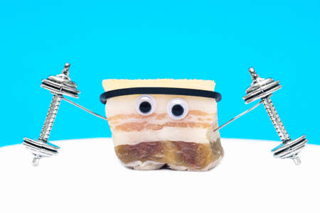 Close-up of a funny character made of salted pork fat lifting dumbbells on a light blue background. Stay fit creative concept.の写真素材