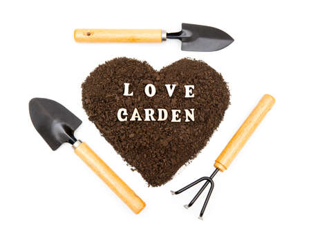 Creative arrangement of garden hand tools around a heart shaped pile of soil with lettering LOVE GARDEN made of wooden characters isolated on white background. Gardening devotion concept.の写真素材