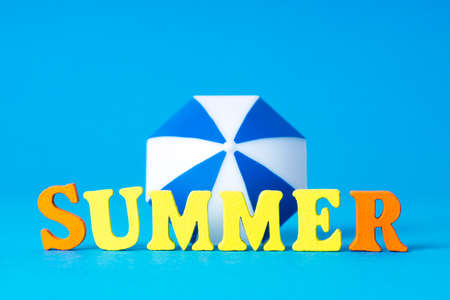 Word SUMMER made of wooden letters and a toy sun umbrella on blue background. Creative sea vacation concept.の写真素材