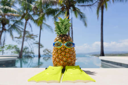 Funny pineapple character wearing goggles and swim fins by a swimming pool on a sunny day against a tropical background. Summer vacation concept.の写真素材