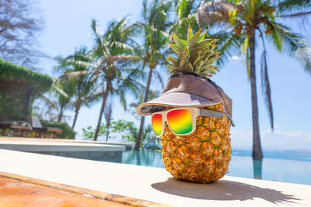 Funny pineapple character wearing gradient sunglasses and a cap by a swimming pool. Tropical vacation destination concept.の写真素材