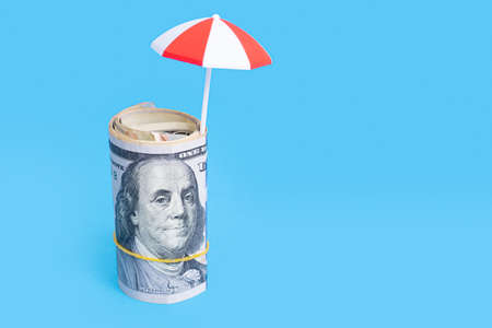 Roll of dollar bills with a toy sun umbrella isolated on blue background. Summer vacation savings concept.の写真素材