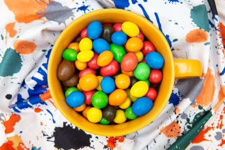 Orange mug full of multicolored candy dragees on a vivid painted cloth backgroundの写真素材