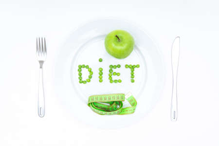 Word DIET arranged of garden peas and served on a large white plate with a fresh green apple, a soft measuring tape, knife and fork isolated on white.の写真素材