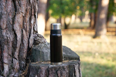 Black vacuum flask on a tree stump. Blurred forest background, copy space.の写真素材