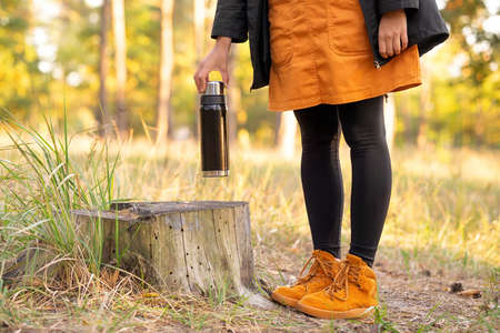 Female tourist wearing orange clothes placing a thermo flask on a tree stump in the forest to have a...