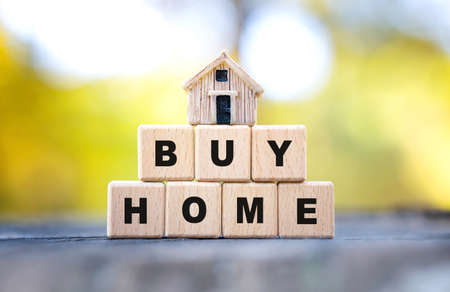 Text BUY HOME made of wooden letter blocks and a tiny toy house standing on a tree stump against a blurred autumn forest background. Countryside dream home concept.