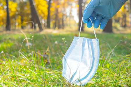 Close-up of a hand wearing a disposable glove picking up a discarded single-use face mask from the...